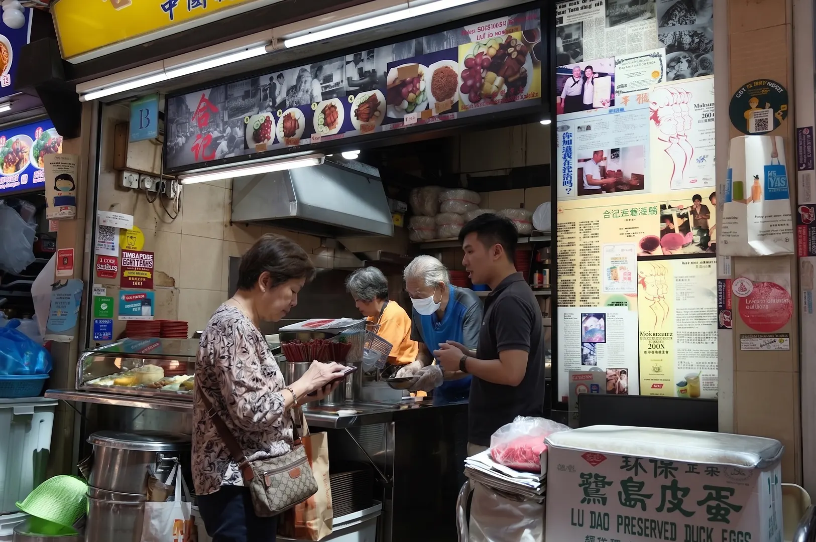 Customers ordering food at the Lu Dao Preserved Duck Eggs stall, a traditional Singapore hawker stall at People's Park Food Centre, featuring elderly vendors and newspaper clippings on the storefront.