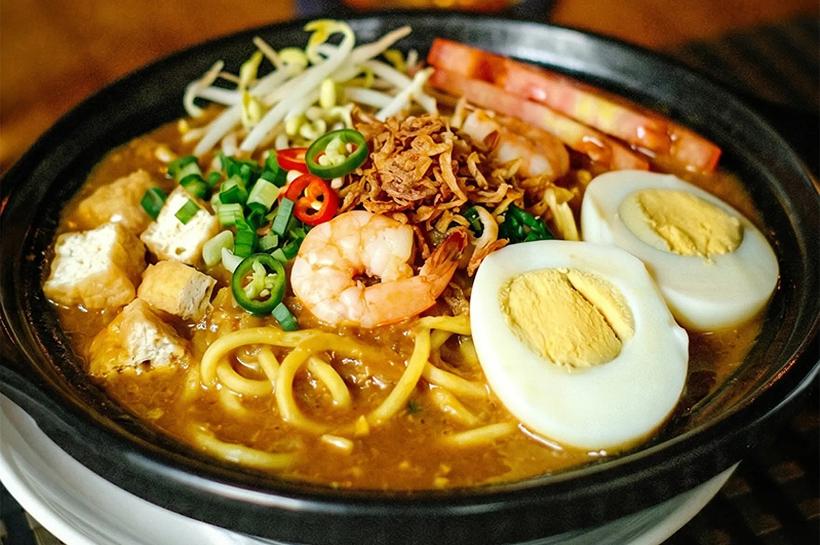 Malaysian Mee Rebus noodle soup with shrimp, hard-boiled egg, fried tofu, and bean sprouts in a black bowl.
