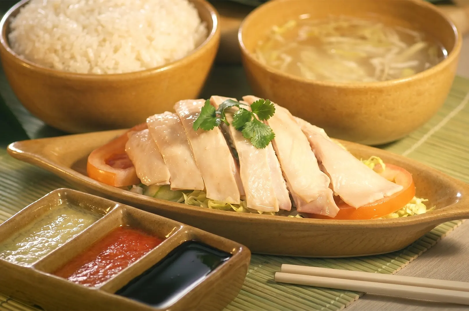 Poached Hainanese chicken served with fragrant rice, clear soup, and a trio of ginger, chili, and dark soy dipping sauces.