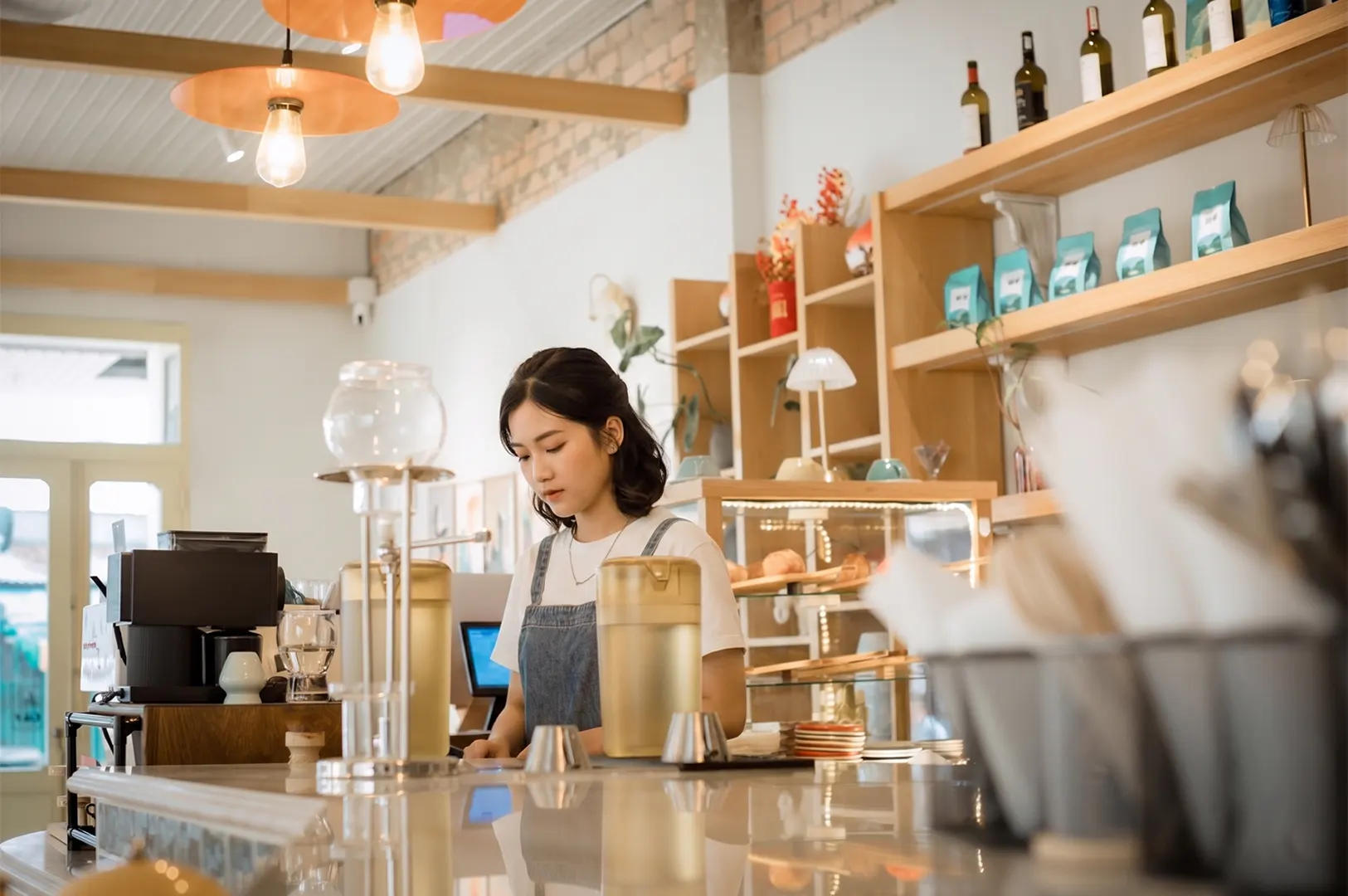 A female barista preparing coffee behind a modern cafe counter with shelves of coffee beans.