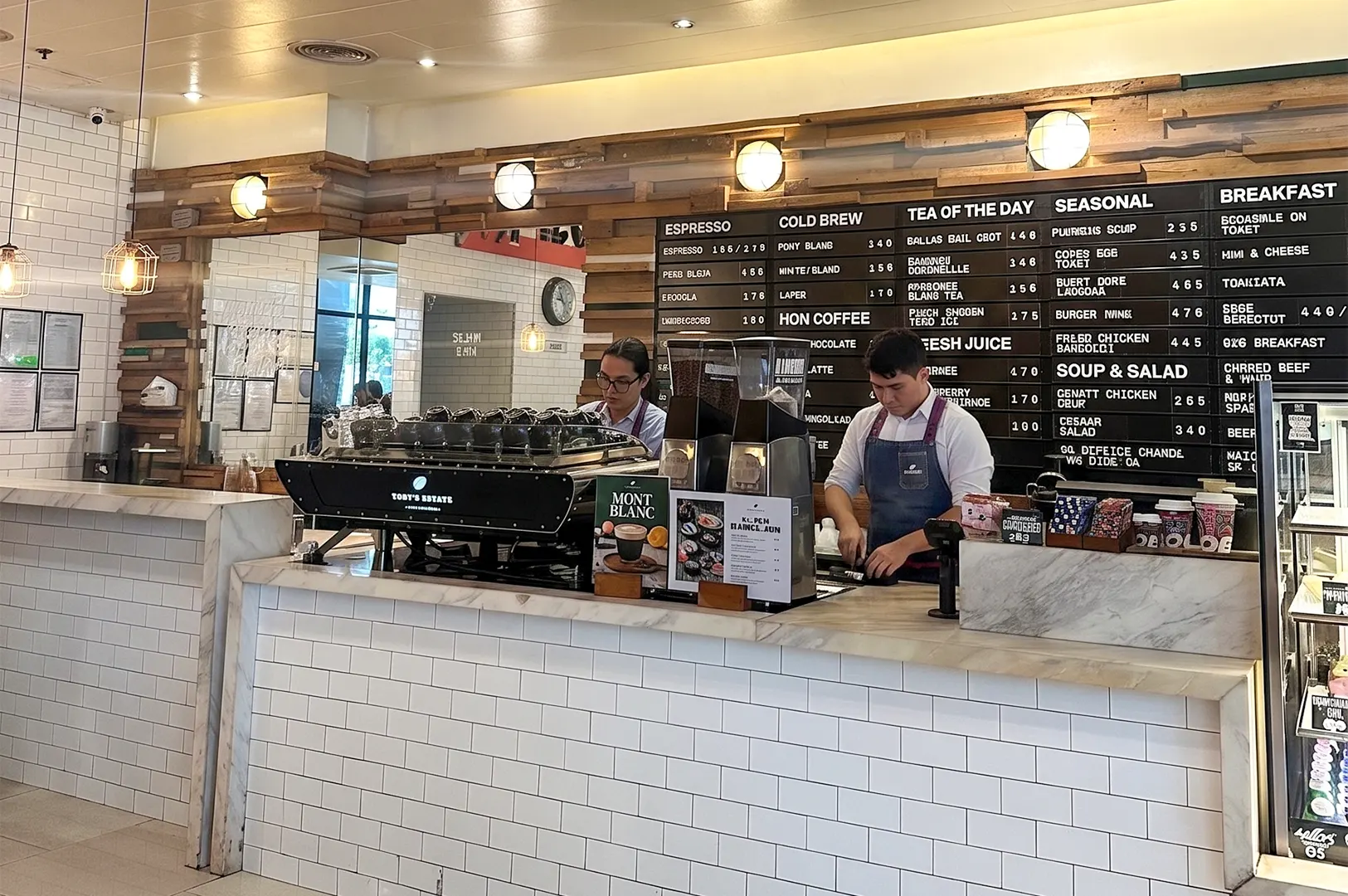 Baristas working at a Toby's Estate coffee shop counter with a large menu board and espresso machine.