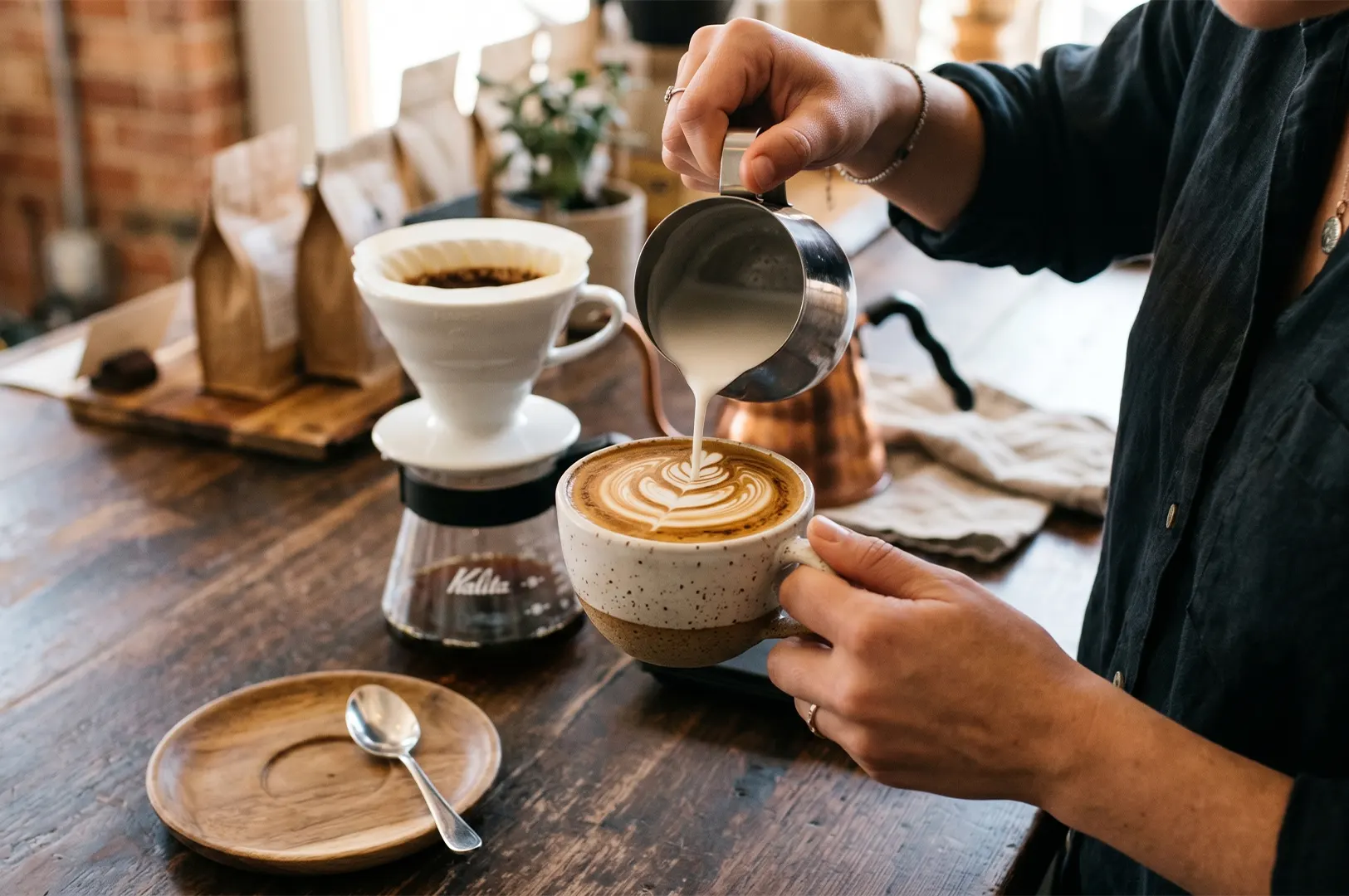 Close-up of a barista pouring steamed milk from a metal pitcher to create latte art in a ceramic cup, with a Kalita pour-over coffee dripper setup on a wooden counter.